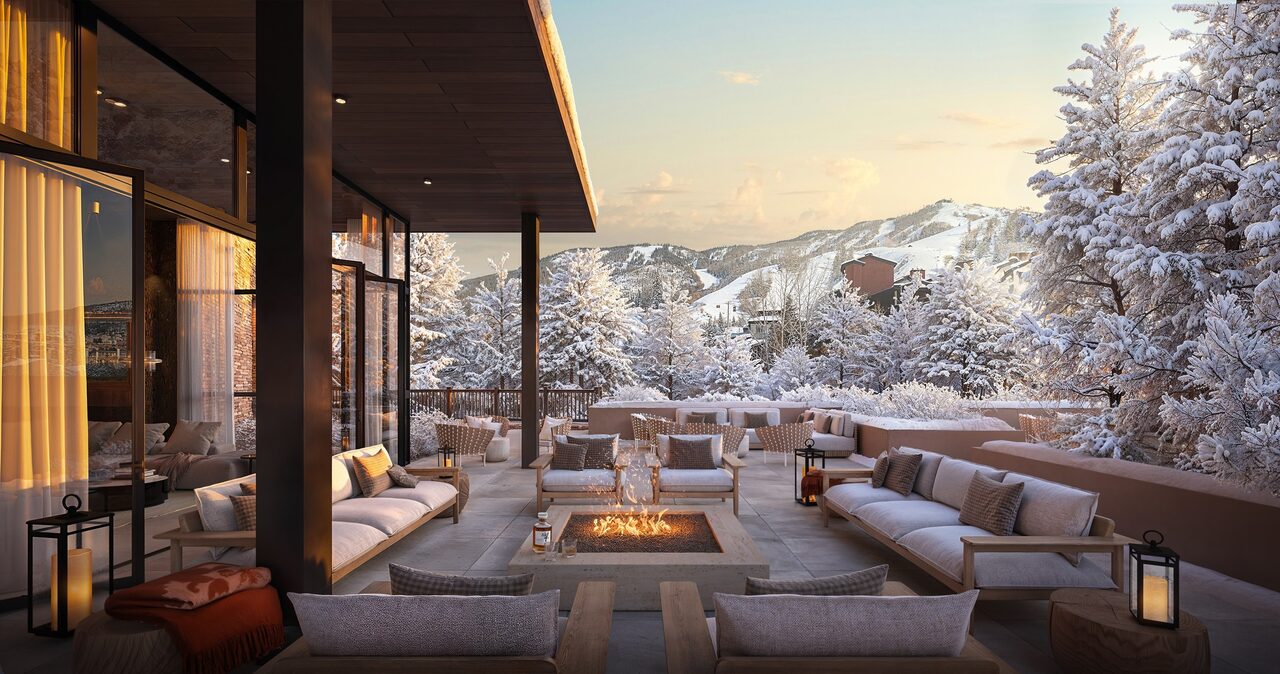 A cozy outdoor lounge with modern seating, a fire pit, and snowy mountain views at sunset. Warm lighting enhances the inviting atmosphere.